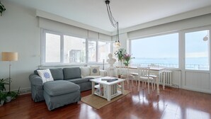 Un ampio soggiorno con divano grigio, tavolino bianco e grandi finestre che offrono vista sul mare.