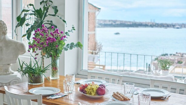 Tavola imbandita con piatti, bicchieri e una ciotola di frutta, con vista su un corpo d'acqua e sullo skyline della città.