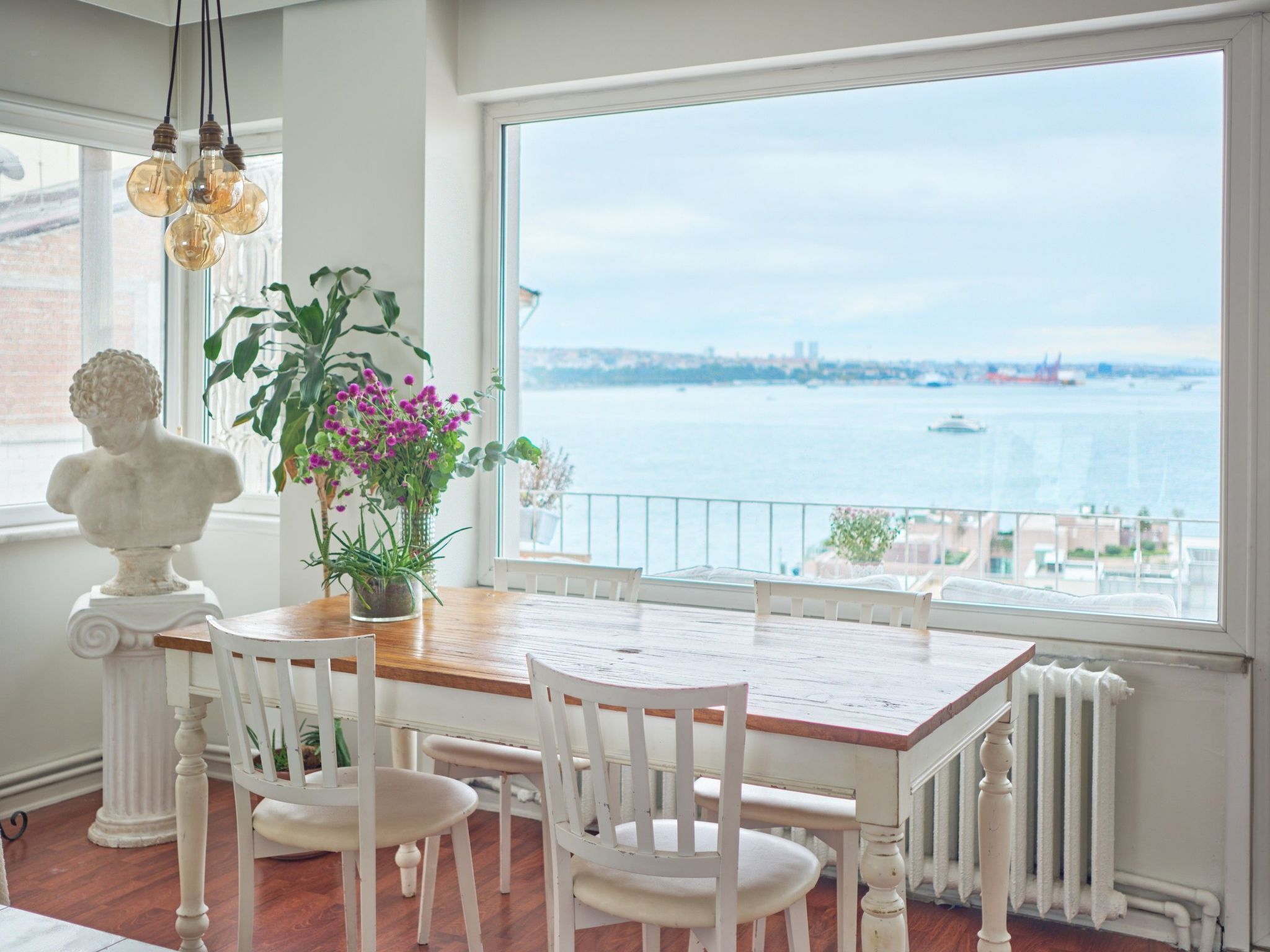 A dining area with a wooden table and white chairs, a potted plant, and a view of the water and city skyline.