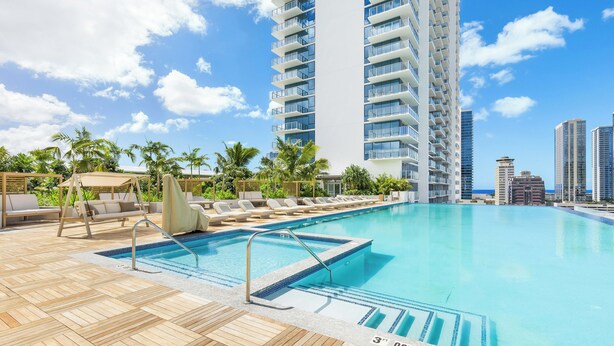 A pool area with lounge chairs and a wooden deck, surrounded by palm trees and tall buildings. - Chic Studio with Skyline Views at The Park on Keeaumoku (Honolulu)
