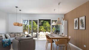 Ein modernes Wohnzimmer mit einem grauen Sofa, einem Esstisch mit Stühlen und Blick durch große Glasüren auf einen Außenbereich.
