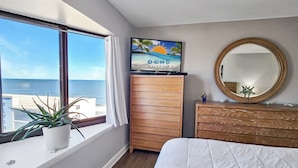 Une chambre avec une grande fenêtre, un lit, une commode en bois, un miroir et un téléviseur diffusant une scène de plage.