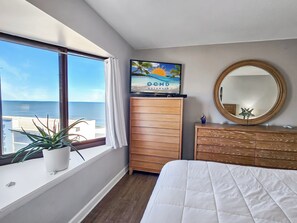 A bedroom with a large window, a bed, a wooden dresser, a mirror, and a television displaying a beach scene. - Direct oceanfront 2 bedroom, 2 bathroom corner condo! 94001304 (Ocean City)