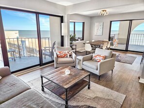 A spacious living room with a large sliding glass door leading to a balcony with a view of the ocean. There are two armchairs, a coffee table, and a dining area with a wooden table and chairs. - Direct oceanfront 2 bedroom, 2 bathroom corner condo! 94001304 (Ocean City)