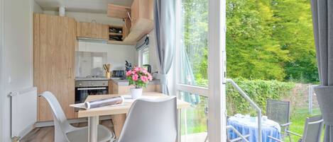 A modern kitchen with wooden cabinets, a dining area with two chairs, a table, and a view of greenery outside.