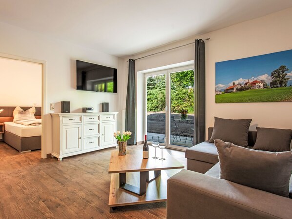 A modern living room with a sofa, a coffee table, a TV mounted on the wall, and a large window with a view of greenery and buildings. - Alpenlodge Hochvogel by Interhome (Lechbruck am See)