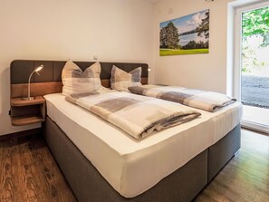 A modern bedroom with a large bed, a nightstand, a lamp, and a window with a view of greenery. - Alpenlodge Hochvogel by Interhome (Lechbruck am See)