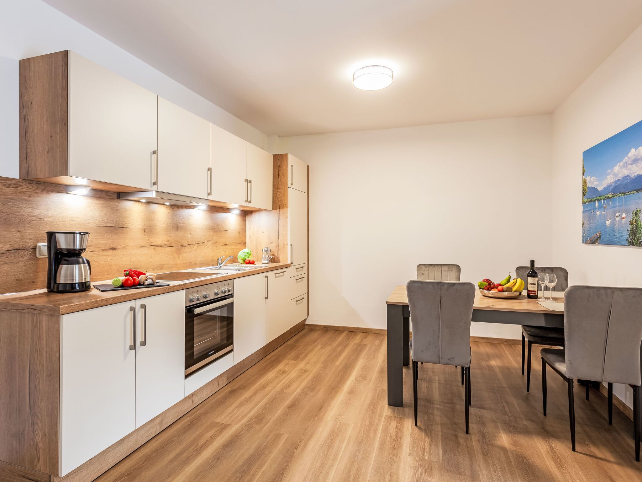 A modern kitchen with wooden cabinets, a built-in oven, and a coffee maker. There is a dining table with chairs and a fruit bowl.
