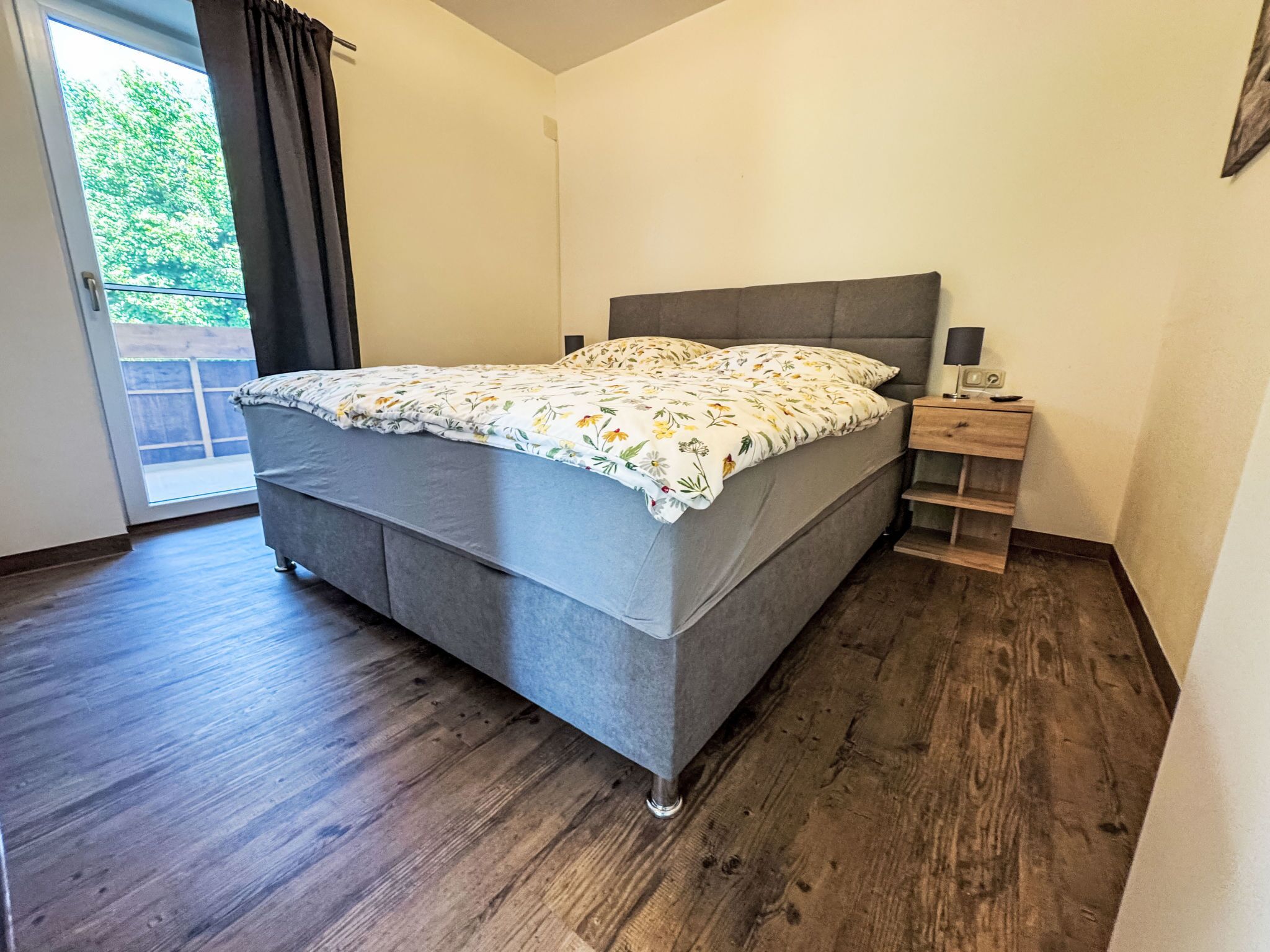 A bedroom with a bed, a nightstand, a lamp, and a view of greenery outside.