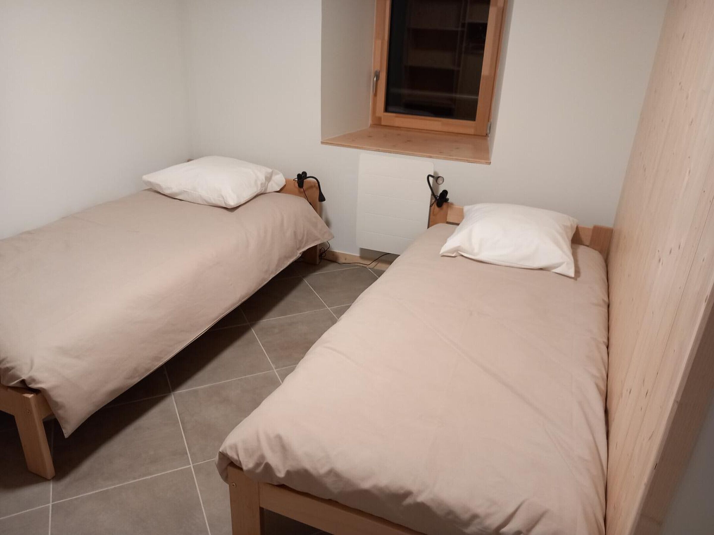 Two single beds with wooden frames and beige bedding, a small window above one bed, and tiled flooring.