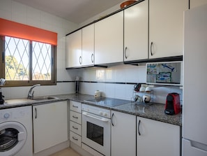 A compact kitchen with white cabinets, a built-in oven, a washing machine, and a window with a view of trees. - 2BR Apartment in Creixell with Pool & Parking, 5min to Beach (Creixell)