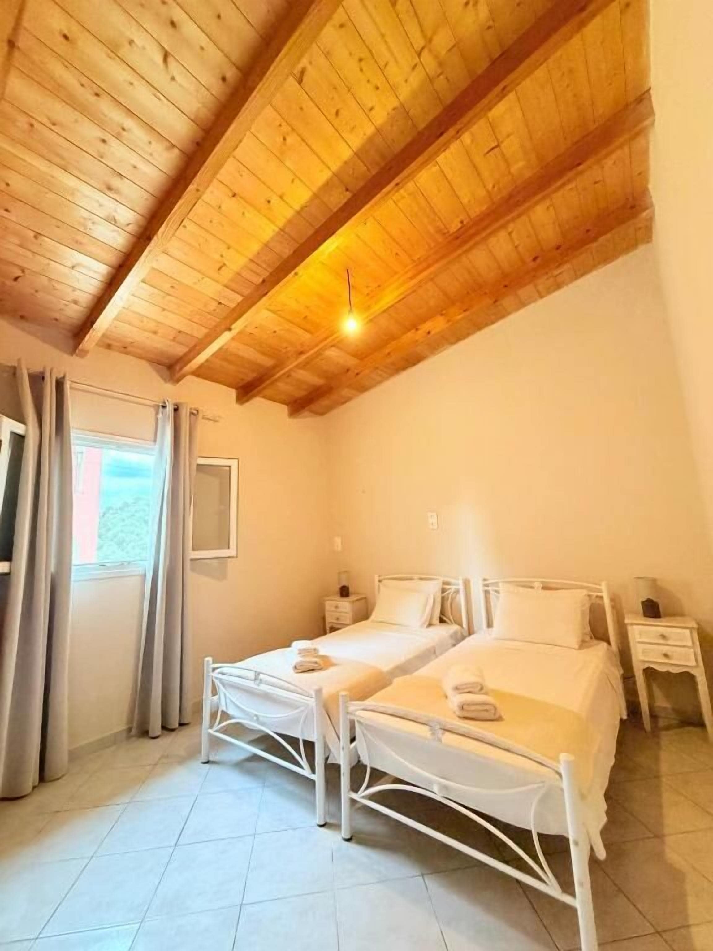 Una stanza con due letti singoli, soffitto in legno, una finestra con tende e un comodino con lampada.