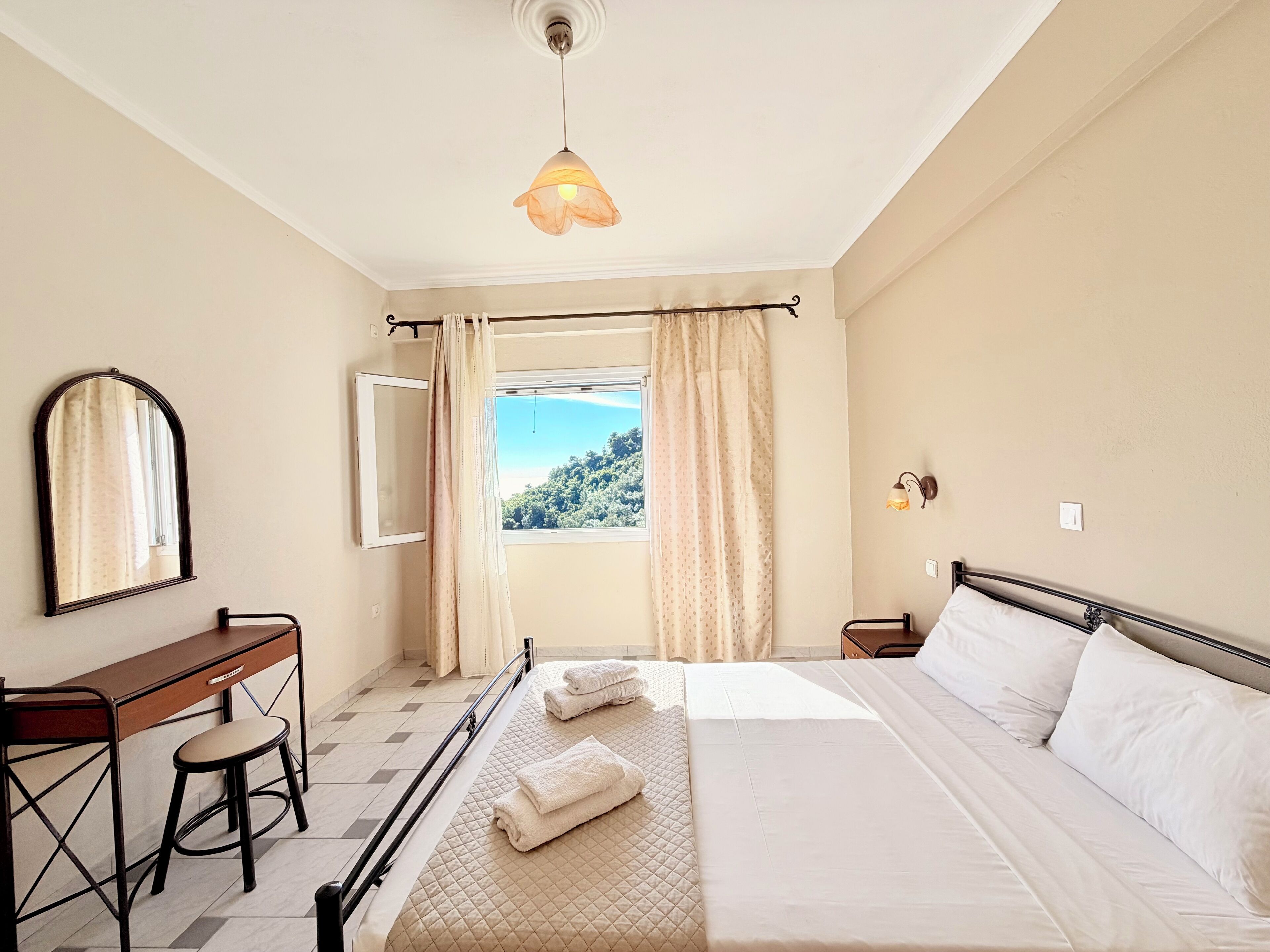 Ein Hotelzimmer mit Bett, Schreibtisch, Stuhl, Spiegel, Fenster mit Vorhängen und einer Pendelleuchte.