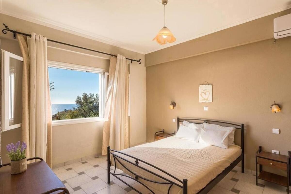 A bedroom with a bed, a nightstand, a bedside table, a window with curtains, and a view of the sea.