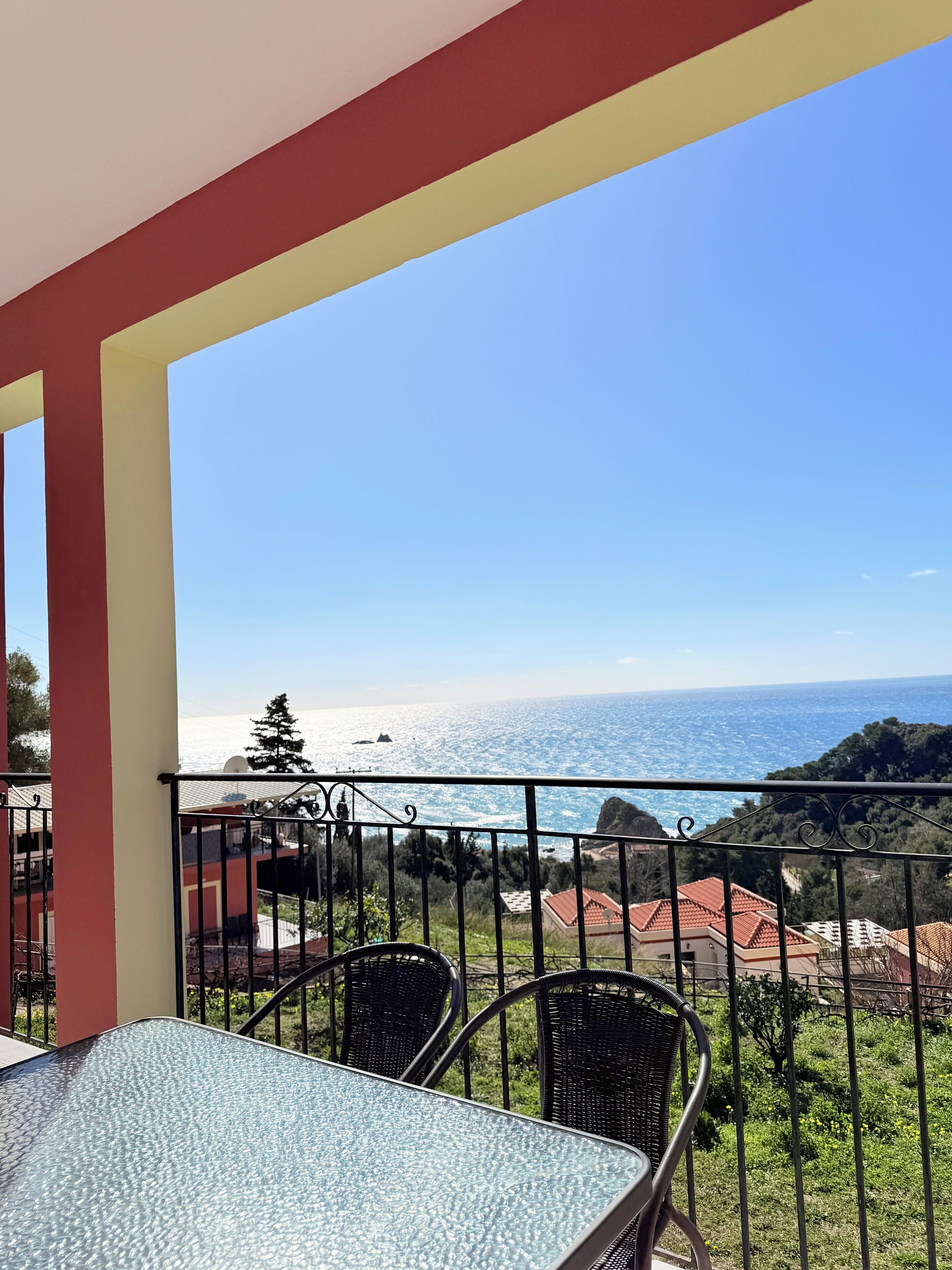 A balcony with a table and chairs, overlooking a coastal view with buildings and greenery.