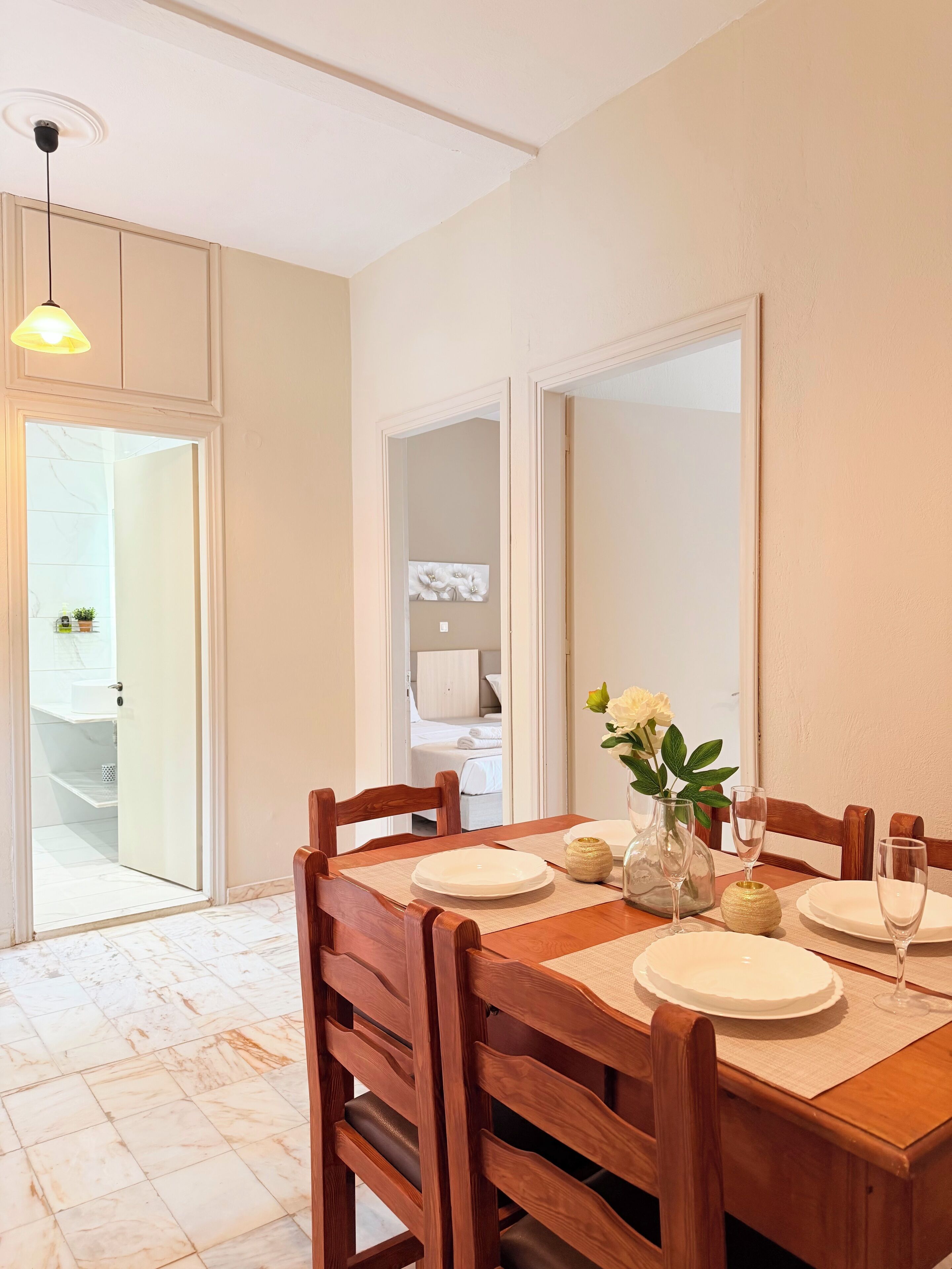 A dining area with a wooden table set for four, a vase with flowers, and a view into a bedroom with a bed and a nightstand.
