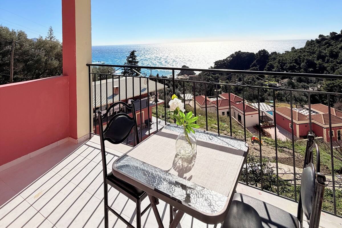 A balcony with a table and chairs, overlooking a coastal view with buildings and trees.