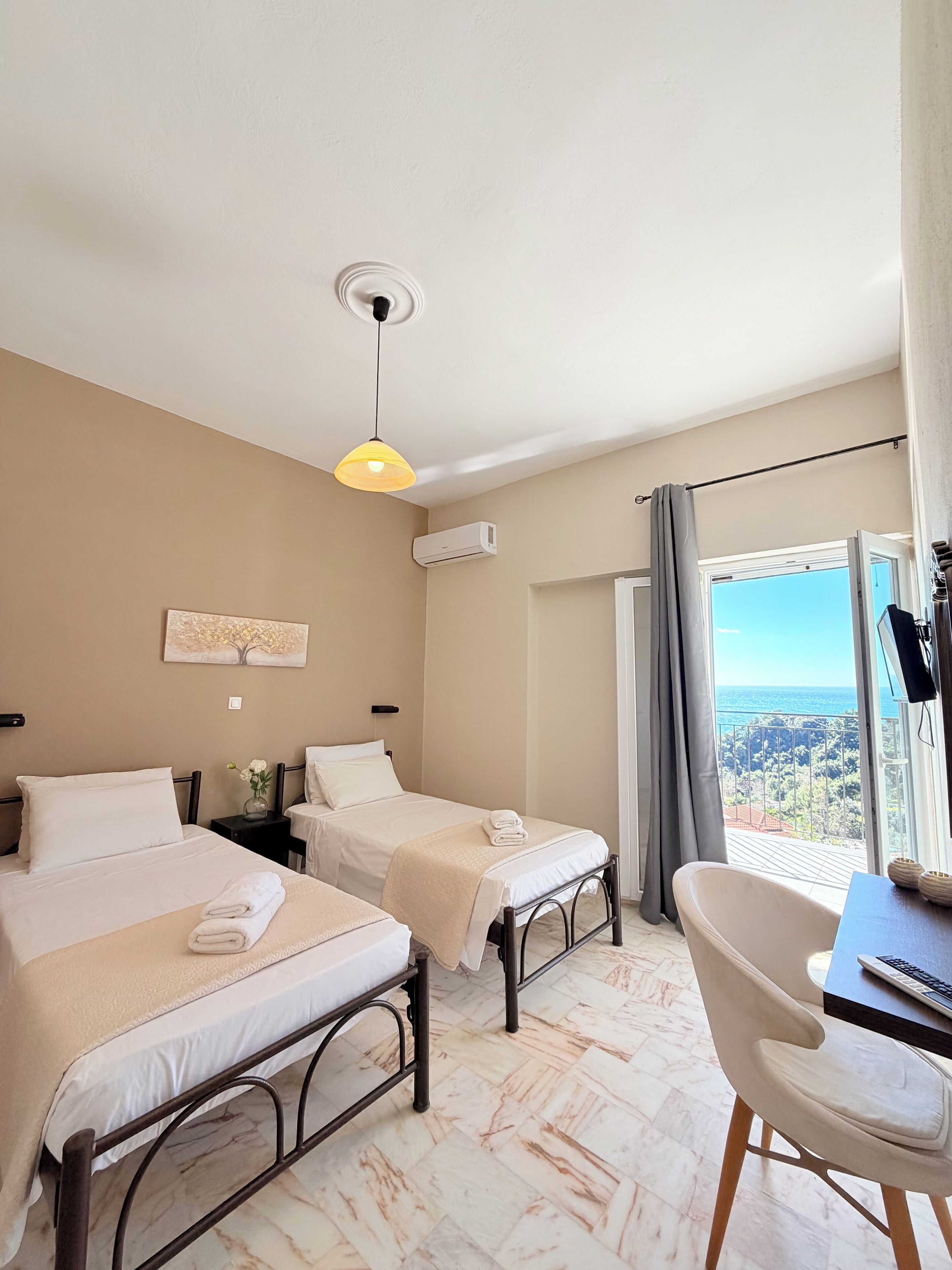 Une chambre d’hôtel avec deux lits, un bureau, une chaise, un appareil de climatisation, un luminaire au plafond et une vue sur la mer.