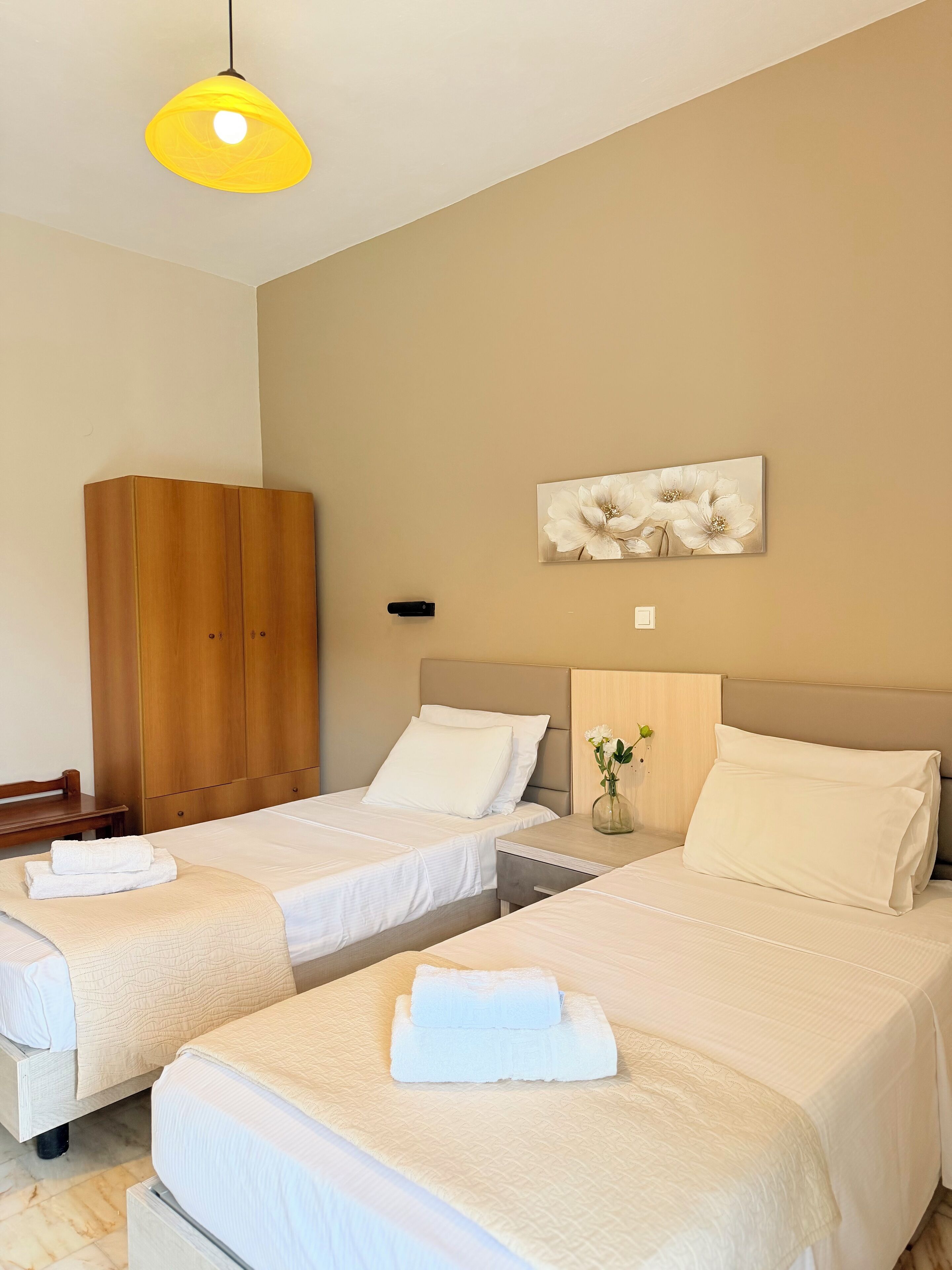 A hotel room with two beds, a wooden wardrobe, a bedside table with a vase, and a painting on the wall.