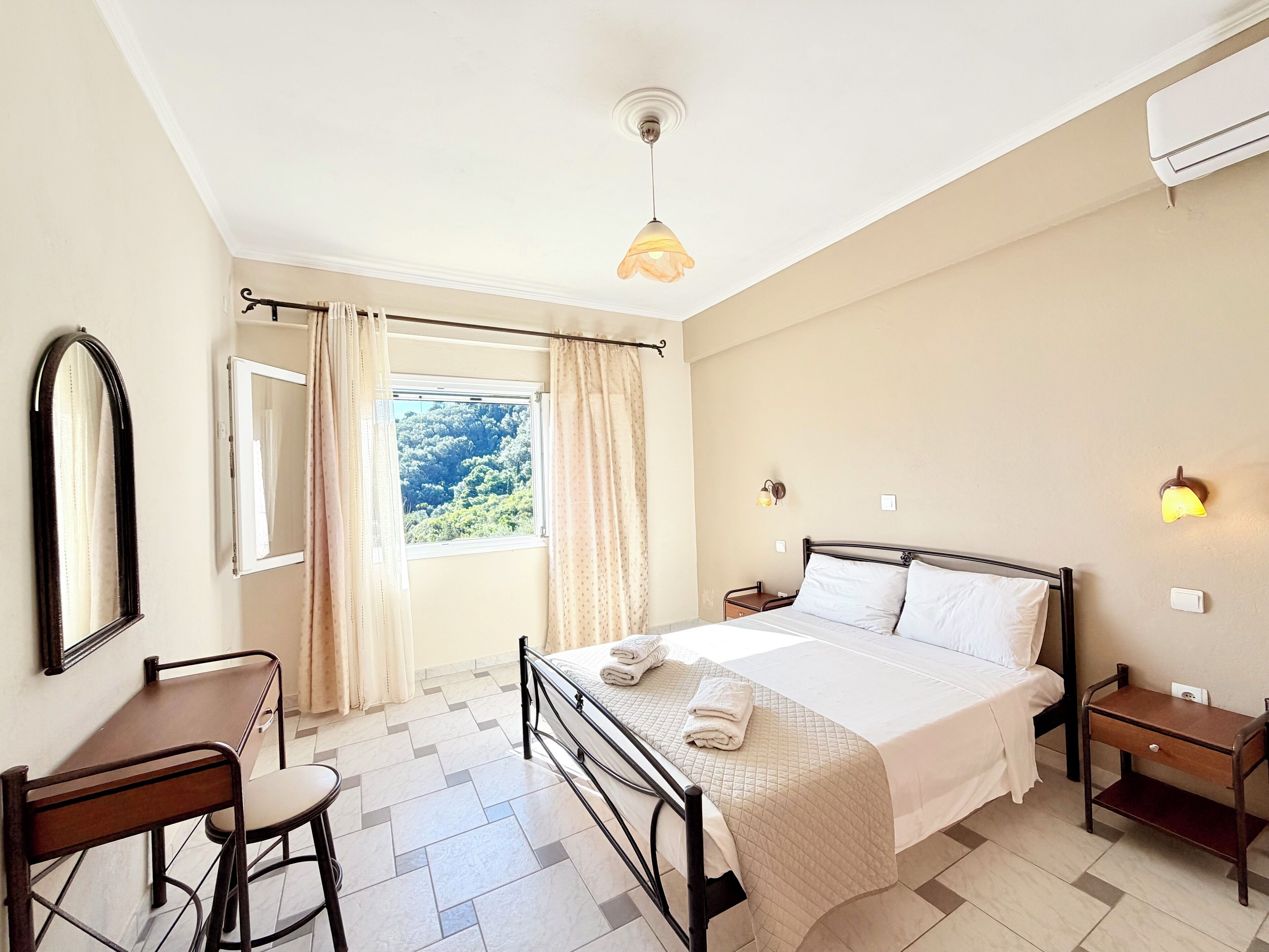A hotel room with a bed, bedside tables, a desk with a chair, a window with curtains, and a view of greenery outside.