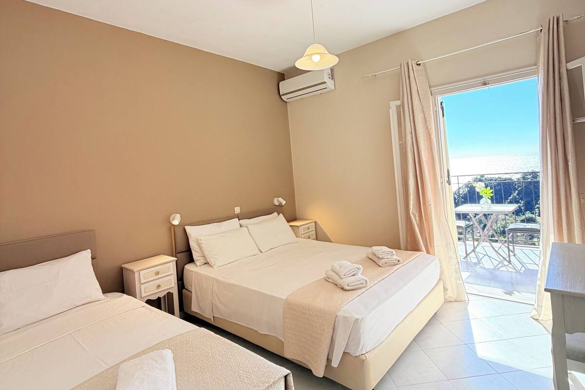 A hotel room with two beds, a ceiling fan, an air conditioning unit, a small table with a plant, and a balcony with a view.