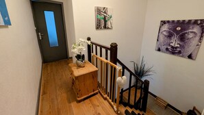 A hallway with wooden flooring, a small wooden bench with a vase of flowers, a staircase with a black railing, and a Buddha statue on the wall. - Spacious Gîte in Selles, 3 Bedrooms, Garden, Family-Friendly (Selles)