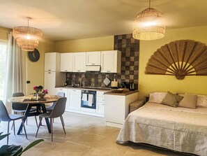 Eine moderne Küche mit weißen Schränken, ein Essbereich mit einem runden Tisch und Stühlen sowie ein Bett mit einer beigen Tagesdecke.
