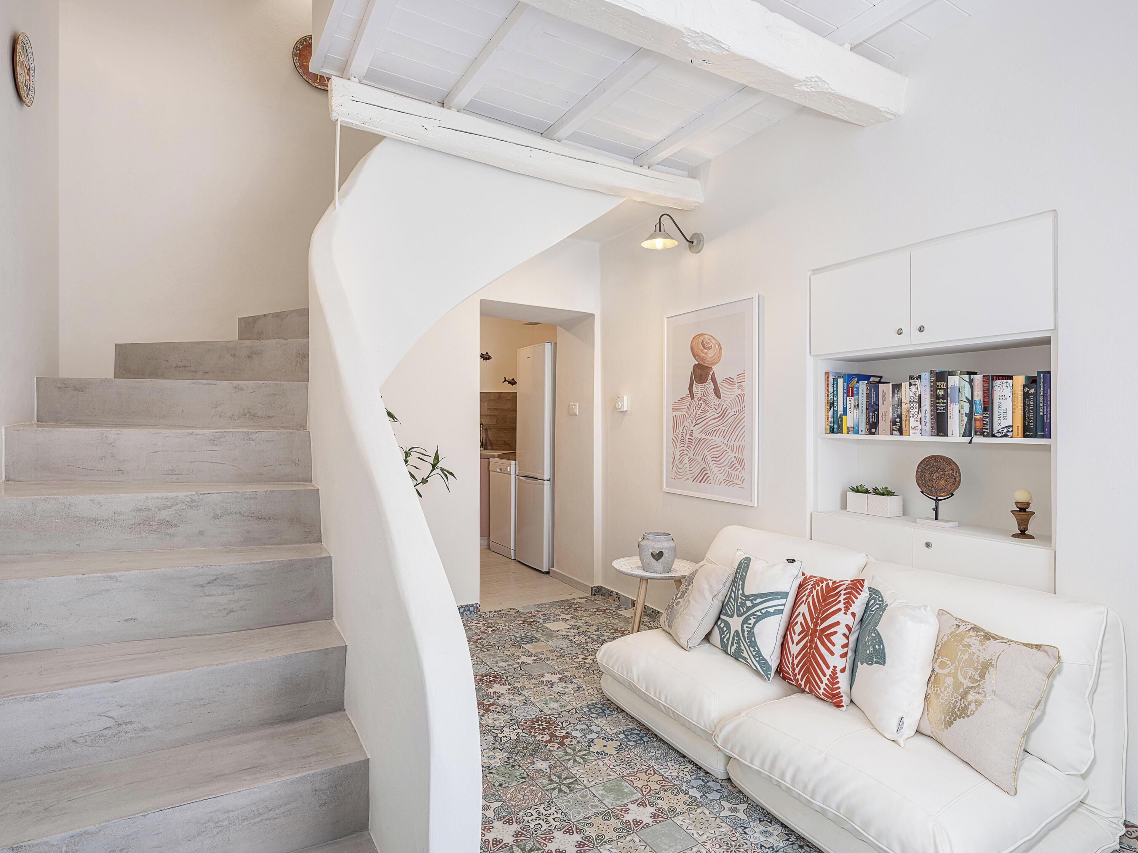 A modern interior with a white staircase, a white sofa with patterned cushions, a kitchen area, and a bookshelf.