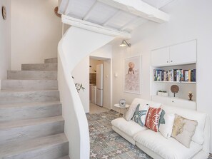 A modern interior with a white staircase, a white sofa with patterned cushions, a kitchen area, and a bookshelf. - Dante e Allegra by Villaspo Homes (Chania)