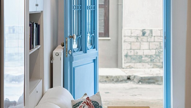 Ein gemütliches Zimmer mit einem beigen Sofa, einem kleinen runden Tisch mit einem Keramiktöpfchen und einer blauen Tür, die nach draußen führt.