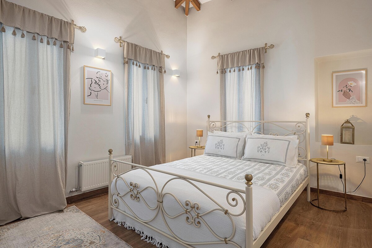 A bedroom with a white metal bed frame, two bedside tables with lamps, a radiator, and a large window with curtains.