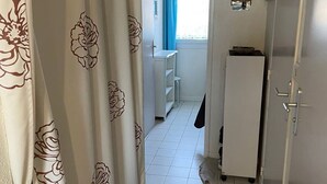 Un pasillo estrecho con una puerta blanca, un pequeño rincón de cocina y un ventanal con cortinas azules.