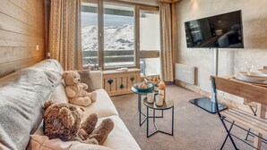 Ett mysigt rum med en soffa, ett stort fönster med utsikt över bergen, en tv och ett litet bord med en flaska och glas.