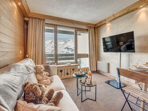 Ein gemütliches Zimmer mit einer Couch, einem großen Fenster mit Blick auf die Berge, einem Fernseher und einem kleinen Tisch mit einer Flasche und Gläsern.