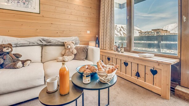 Ett mysigt rum med en väggpanel av trä, ett stort fönster med utsikt över ett snötäckt landskap, en soffa med två nallebjörnar, ett litet bord med croissanter och en flaska juice samt en inramad tavla på väggen.
