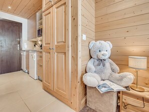 Une pièce chaleureuse aux murs en bois, avec un grand ours en peluche assis sur un petit tabouret. Une lampe et une télécommande sont posées sur une petite table d’appoint en bois.