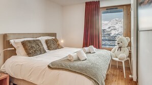 Ein Schlafzimmer mit einem Bett, Kissen, einer Decke, einem Teddybären und Blick auf einen verschneiten Berg durch ein Fenster.