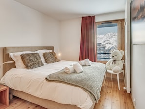 Ein Schlafzimmer mit einem Bett, Kissen, einer Decke, einem Teddybären und Blick auf einen verschneiten Berg durch ein Fenster.