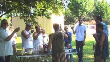 A group of people gathered around a table with food and drinks, outdoors under a tree.