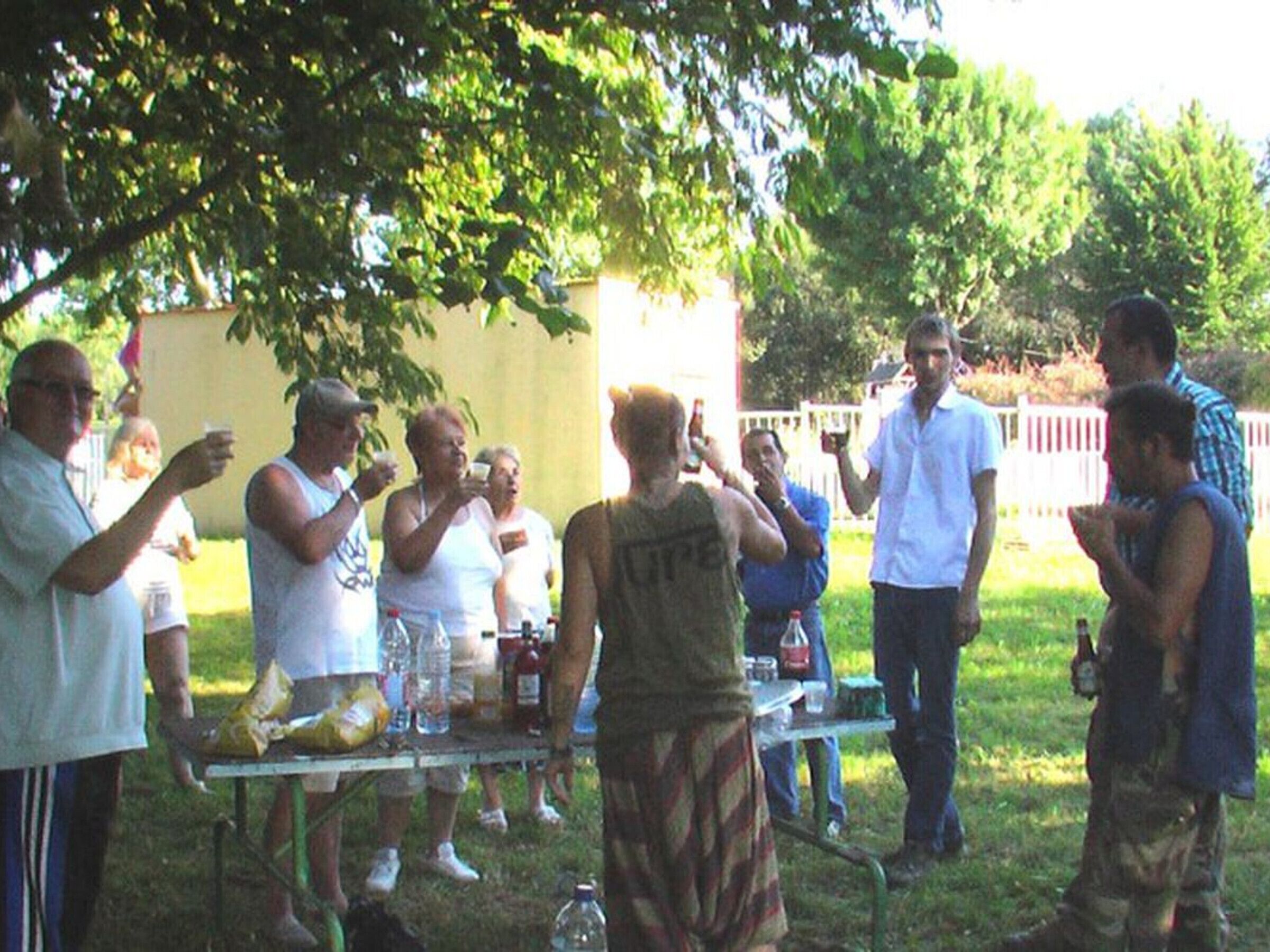 Un grupo de personas reunidas alrededor de una mesa con comida y bebidas, al aire libre bajo un árbol.