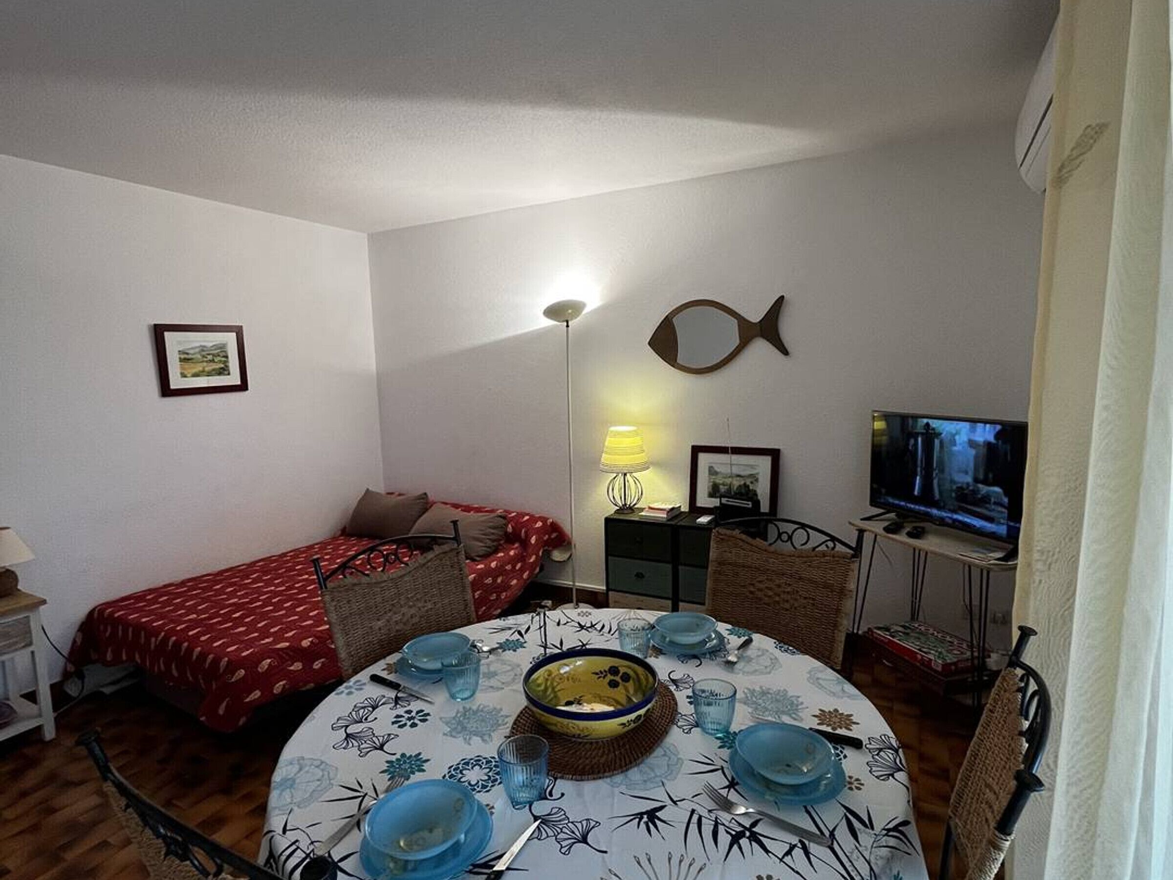 A compact room with a bed, a dining table set with blue dishes, a fish-shaped wall decoration, a television, and a lamp.