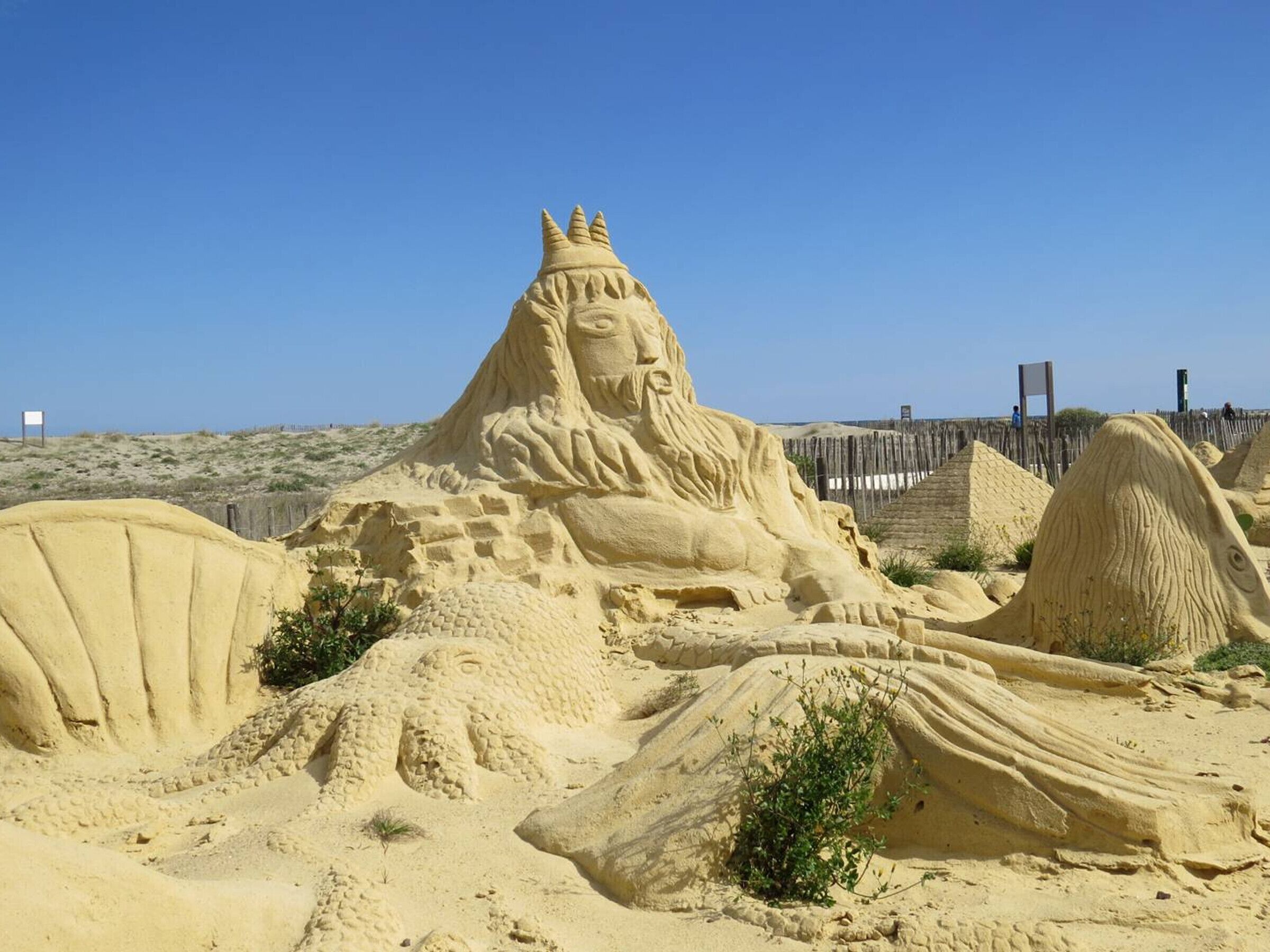 Escultura de areia representando uma figura com coroa e barba, cercada por montes de areia e vegetação rala.