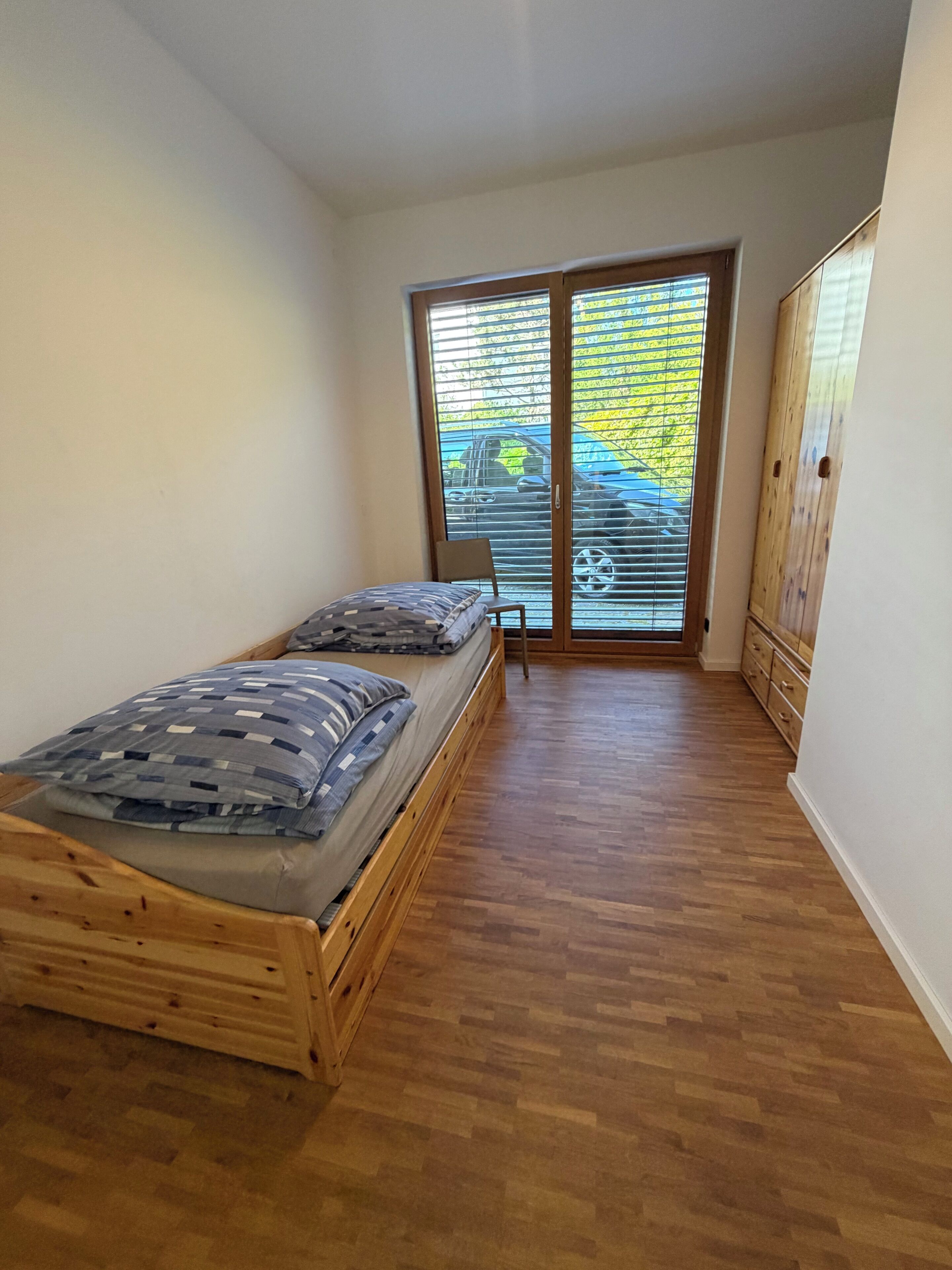 Une chambre à coucher avec un lit en bois, une chaise, une armoire et une porte-fenêtre coulissante donnant sur un balcon.