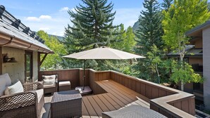 Um deck de madeira com móveis de vime, um guarda-sol grande e árvores altas ao fundo.