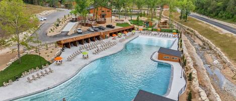 A large outdoor swimming pool with lounge chairs and umbrellas, surrounded by buildings and a hilly landscape.