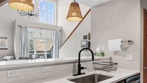 A modern kitchen with white countertops, a stainless steel sink, wicker pendant lights, and a window with curtains. - Spacious Family-Friendly Condo With Updated Finishes (Frisco)