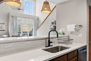 A modern kitchen with white countertops, a stainless steel sink, wicker pendant lights, and a window with curtains. - Spacious Family-Friendly Condo With Updated Finishes (Frisco)