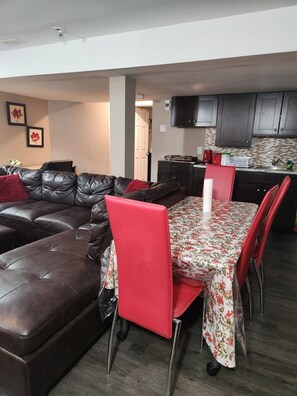 A modern living room with a brown leather sectional sofa, a red chair, and a dining table with a floral tablecloth. - 2BR luxe Lower Level Seperate Entrance (Gatineau)