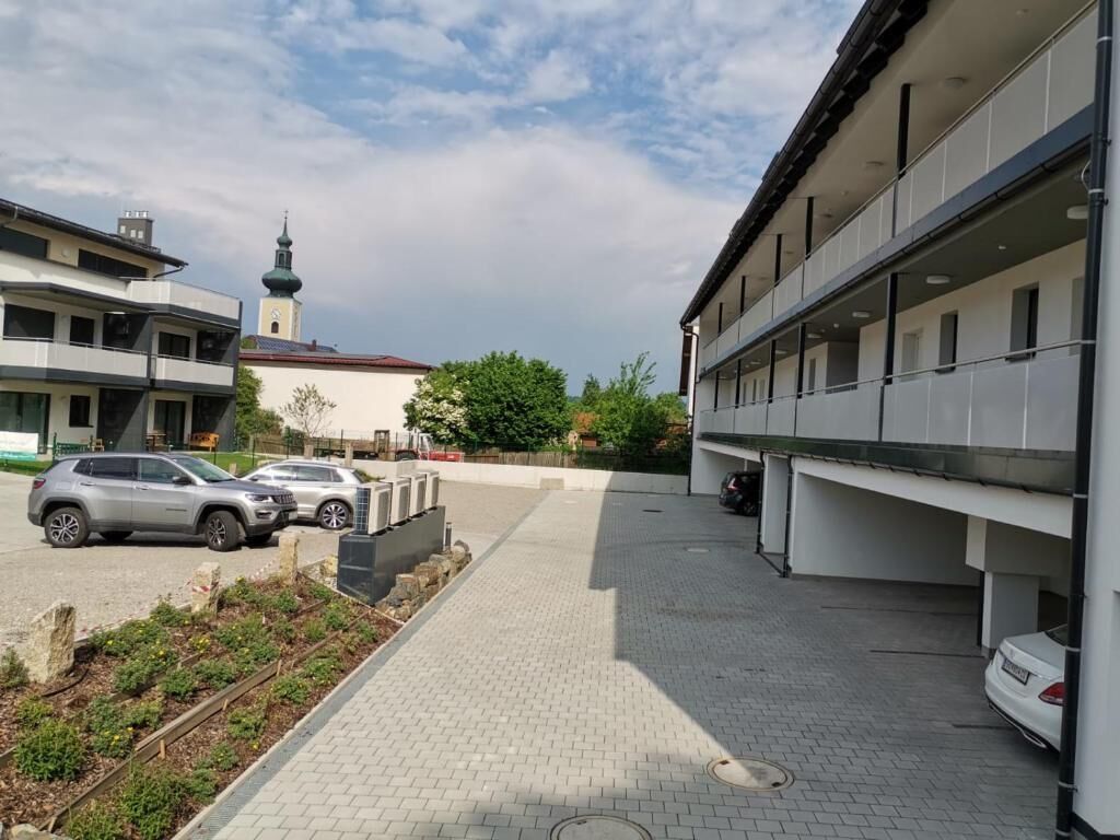 A parking area with cars, a building with a modern design, and a church tower in the background.