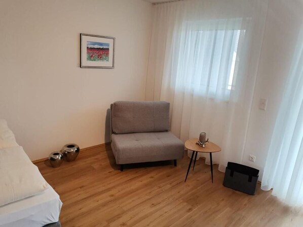 A room with a grey sofa, a small round table with a lamp, a framed picture on the wall, and a black storage box.